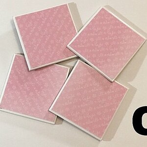 May include: Four pink square coasters with a white script pattern. The coasters are stacked on top of each other.