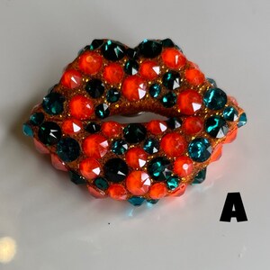 May include: A bejeweled lips-shaped brooch. The brooch features a brown base covered in orange, teal, and clear rhinestones. The letter "A" is visible in the lower right corner.