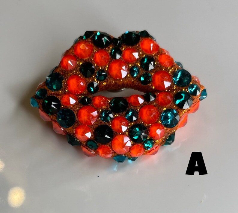 May include: A bejeweled lips-shaped brooch. The brooch features a brown base covered in orange, teal, and clear rhinestones. The letter "A" is visible in the lower right corner.