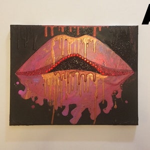 May include: A rectangular canvas painting on a black background featuring a pair of stylized lips in shades of pink and purple. Gold paint drips downward from the upper lip, while a line of small red beads accents the inner edge of the mouth.
