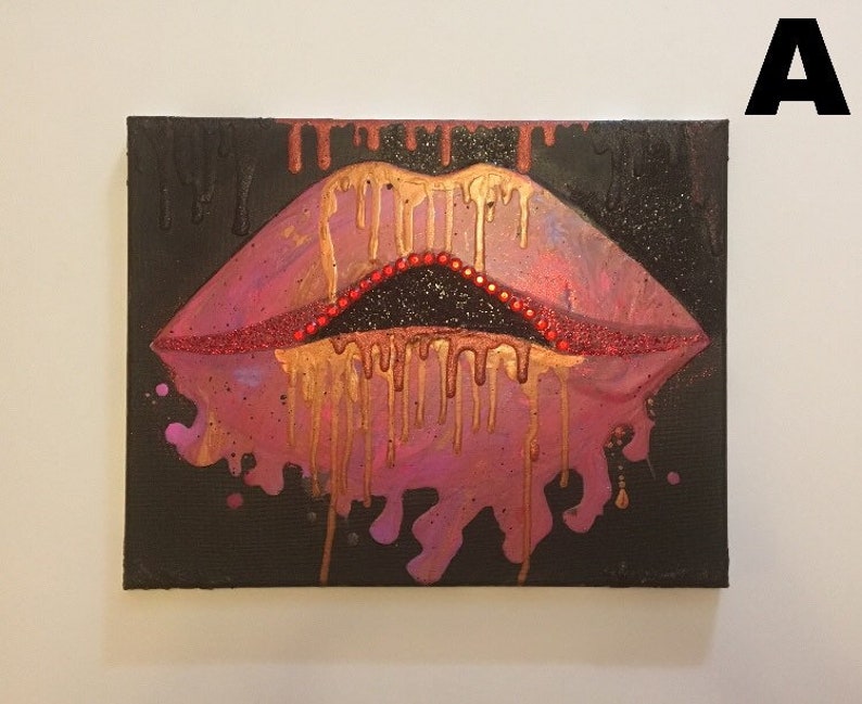 May include: A rectangular canvas painting on a black background featuring a pair of stylized lips in shades of pink and purple. Gold paint drips downward from the upper lip, while a line of small red beads accents the inner edge of the mouth.