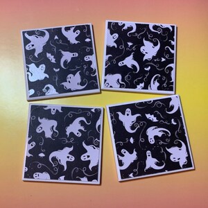 May include: A set of four square coasters featuring a black background with a repeating pattern of white ghosts, small bats, and swirling line accents. Each coaster has a clean white border framing the graphic design on the top surface.