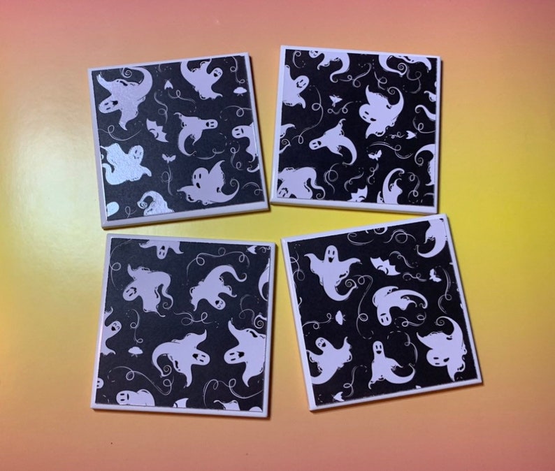May include: A set of four square coasters featuring a black background with a repeating pattern of white ghosts, small bats, and swirling line accents. Each coaster has a clean white border framing the graphic design on the top surface.