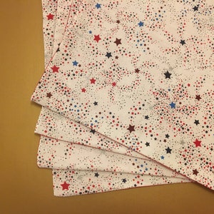 Peut inclure: Quatre serviettes en tissu blanc avec un motif d'&eacute;toiles rouges, bleues et argent&eacute;es. Les serviettes sont pli&eacute;es en pile.