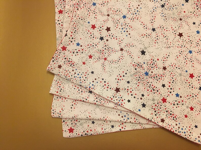Peut inclure: Quatre serviettes en tissu blanc avec un motif d'&eacute;toiles rouges, bleues et argent&eacute;es. Les serviettes sont pli&eacute;es en pile.