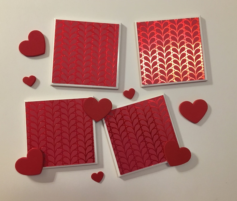 May include: Four red square coasters with a white border and a repeating pattern of red hearts. The coasters are surrounded by red hearts of various sizes.