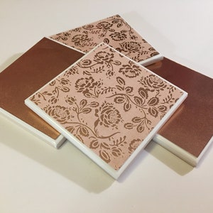 Puede incluir: Conjunto de cuatro posavasos cuadrados con borde blanco y dise&ntilde;o floral rosa y dorado. Dos de los posavasos tienen un color cobre s&oacute;lido.