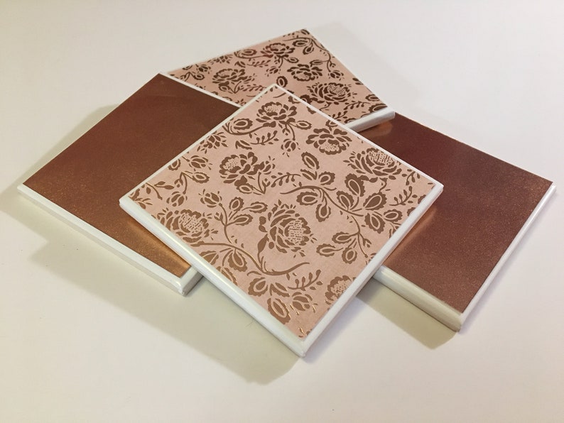 Puede incluir: Conjunto de cuatro posavasos cuadrados con borde blanco y dise&ntilde;o floral rosa y dorado. Dos de los posavasos tienen un color cobre s&oacute;lido.