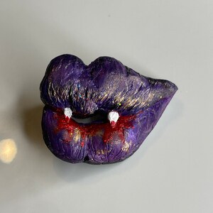 K&ouml;nnte beinhalten: Lila und goldene Glitzerlippen mit wei&szlig;en Z&auml;hnen und rotem, tropfendem Blut. Halloween-Skulptur aus Polymer Clay.