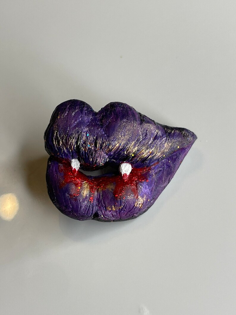 K&ouml;nnte beinhalten: Lila und goldene Glitzerlippen mit wei&szlig;en Z&auml;hnen und rotem, tropfendem Blut. Halloween-Skulptur aus Polymer Clay.