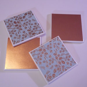 Puede incluir: Cuatro posavasos cuadrados de cer&aacute;mica con un acabado met&aacute;lico de cobre. Dos posavasos presentan un fondo azul claro con un patr&oacute;n floral de cobre. Dos posavasos son de cobre s&oacute;lido.