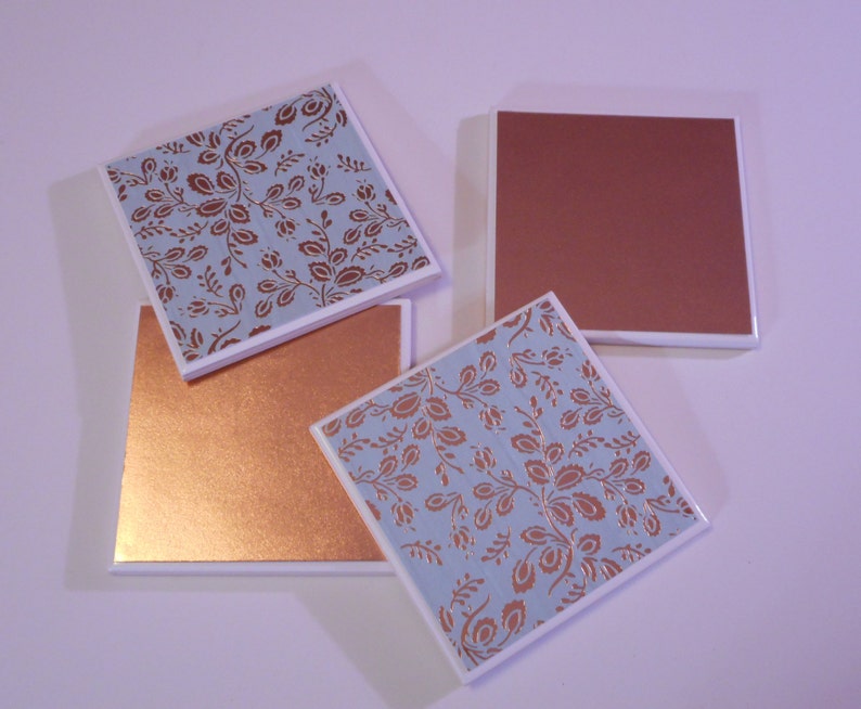 Puede incluir: Cuatro posavasos cuadrados de cer&aacute;mica con un acabado met&aacute;lico de cobre. Dos posavasos presentan un fondo azul claro con un patr&oacute;n floral de cobre. Dos posavasos son de cobre s&oacute;lido.