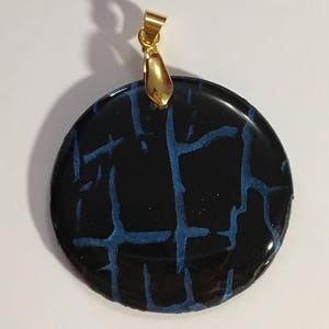Puede incluir: Un colgante redondo con fondo negro y un patr&oacute;n de craquelado azul. El colgante tiene una anilla de oro.