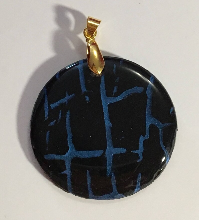 Puede incluir: Un colgante redondo con fondo negro y un patr&oacute;n de craquelado azul. El colgante tiene una anilla de oro.