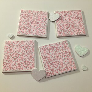 May include: Four white ceramic coasters with a pink and white heart pattern. The coasters are square and have a white border. There are five white glitter hearts scattered around the coasters.