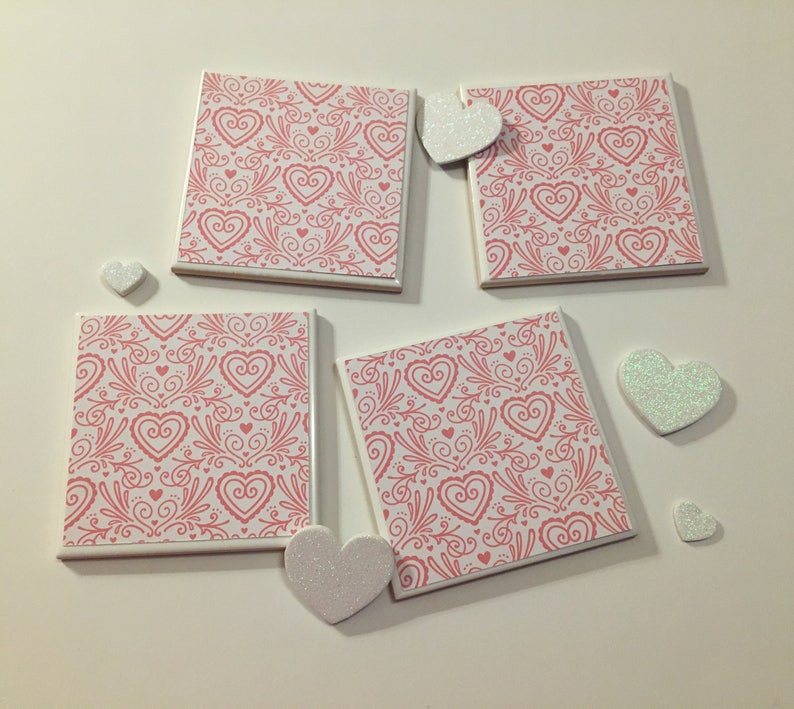 May include: Four white ceramic coasters with a pink and white heart pattern. The coasters are square and have a white border. There are five white glitter hearts scattered around the coasters.