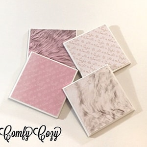 May include: Four square coasters with different designs. Two coasters feature a pink and white faux fur pattern. One coaster has a pink background with a white pattern of small flowers. The last coaster has a white background with a pink pattern of small flowers. The coasters are arranged in a pile on a white surface. The text "Comfy Cozy" is written in black cursive below the coasters.