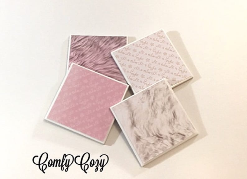 May include: Four square coasters with different designs. Two coasters feature a pink and white faux fur pattern. One coaster has a pink background with a white pattern of small flowers. The last coaster has a white background with a pink pattern of small flowers. The coasters are arranged in a pile on a white surface. The text "Comfy Cozy" is written in black cursive below the coasters.