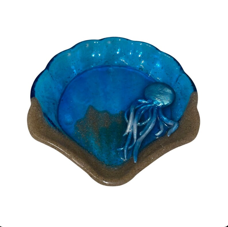 Puede incluir: Un plato de vidrio azul en forma de concha de mar con una figura de pulpo blanca y plateada en su interior. El plato tiene un borde marr&oacute;n arenoso.