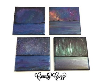 May include: Set of four square canvas paintings depicting different night sky scenes. Each painting features a dark blue water scene with a black horizon line and a colorful night sky above. The skies feature a galaxy, stars, and the aurora borealis.