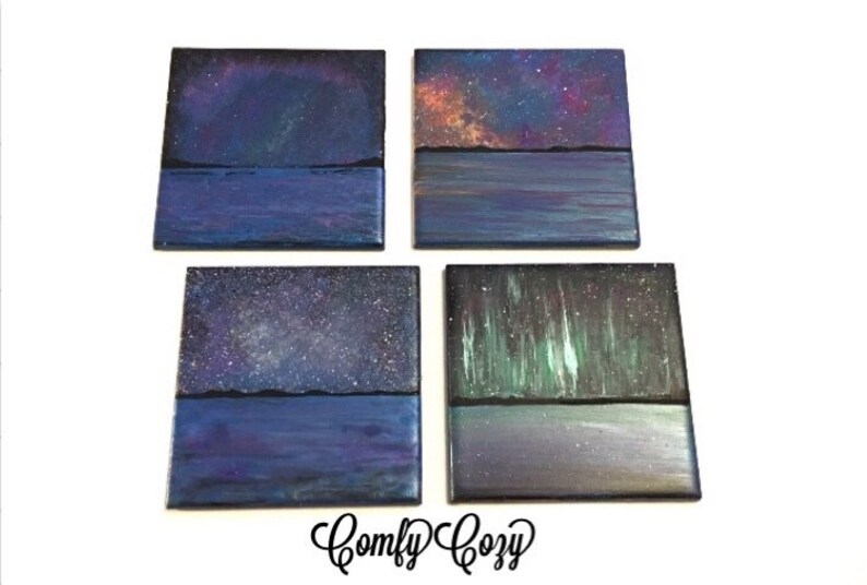 Puede incluir: Conjunto de cuatro pinturas cuadradas sobre lienzo que representan diferentes escenas de cielo nocturno. Cada pintura presenta una escena de agua azul oscuro con una l&iacute;nea de horizonte negra y un cielo nocturno colorido arriba. Los cielos presentan una galaxia, estrellas y la aurora boreal.