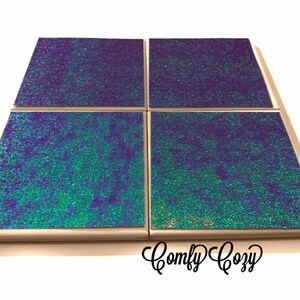 May include: Set of four square coasters with a iridescent green and purple glitter finish. The coasters have a light wood frame and are arranged in a 2x2 grid. The text "Comfy Cozy" is visible in the bottom right corner of the image.