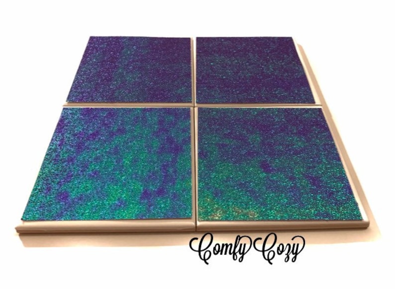 May include: Set of four square coasters with a iridescent green and purple glitter finish. The coasters have a light wood frame and are arranged in a 2x2 grid. The text "Comfy Cozy" is visible in the bottom right corner of the image.