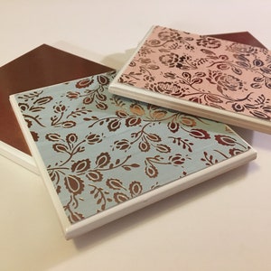 Puede incluir: Tres posavasos cuadrados de cer&aacute;mica con borde blanco. Los posavasos presentan un patr&oacute;n floral en marr&oacute;n y dorado sobre un fondo azul claro, rosa y marr&oacute;n.