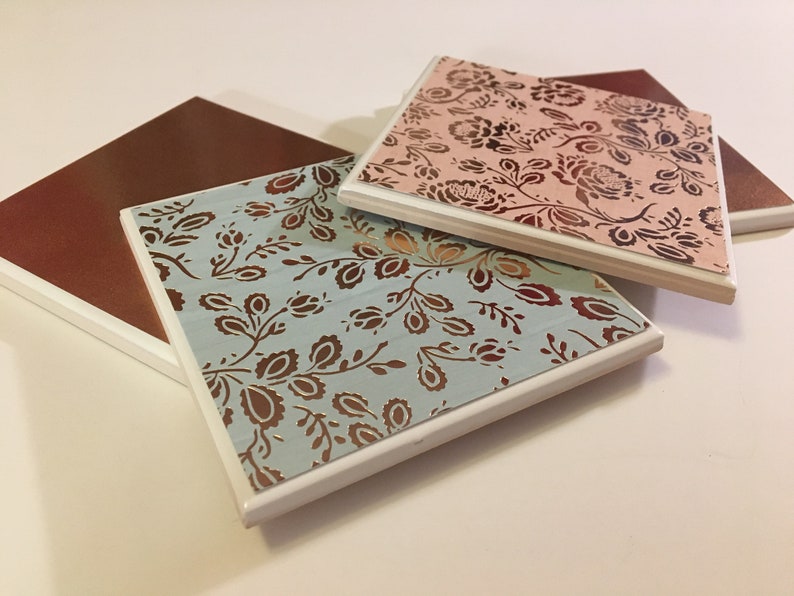 Puede incluir: Tres posavasos cuadrados de cer&aacute;mica con borde blanco. Los posavasos presentan un patr&oacute;n floral en marr&oacute;n y dorado sobre un fondo azul claro, rosa y marr&oacute;n.