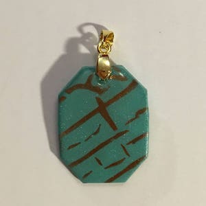 Puede incluir: Un colgante azul turquesa con una anilla de oro y una forma geom&eacute;trica. El colgante tiene un dise&ntilde;o marr&oacute;n y dorado.