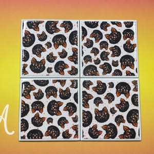 May include: A set of four square coasters featuring a repeating pattern of black cat faces with orange ears and eyes on a white background. The letter A is visible in the bottom left corner. Each coaster has a glossy finish and white edges.