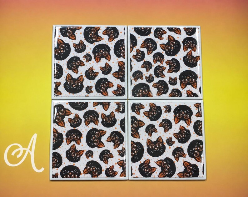 May include: A set of four square coasters featuring a repeating pattern of black cat faces with orange ears and eyes on a white background. The letter A is visible in the bottom left corner. Each coaster has a glossy finish and white edges.