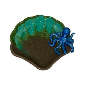 Puede incluir: Un plato en forma de concha de color verde y marr&oacute;n con una figurita de pulpo azul con purpurina en la parte superior. El plato est&aacute; hecho de resina y tiene un acabado brillante.