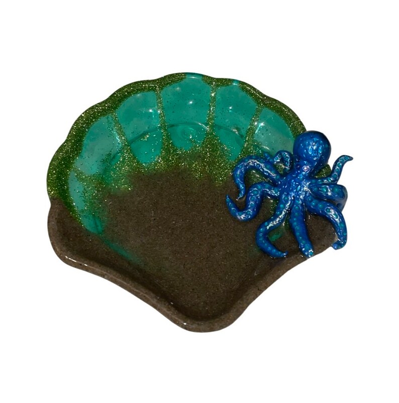 Puede incluir: Un plato en forma de concha de color verde y marr&oacute;n con una figurita de pulpo azul con purpurina en la parte superior. El plato est&aacute; hecho de resina y tiene un acabado brillante.