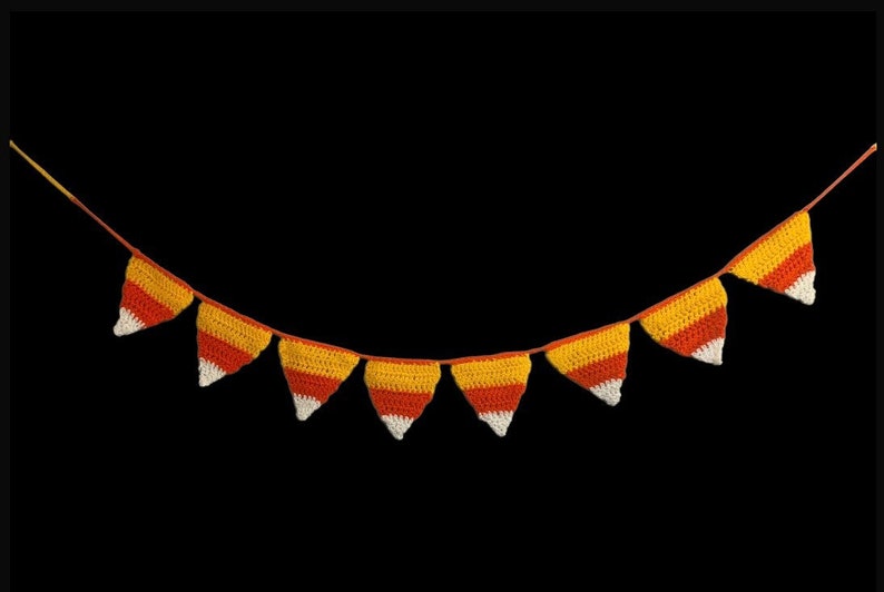 May include: A crocheted garland of triangular pennants in the colors of candy corn: white, orange, and yellow. The pennants are strung on a thin, orange string, perfect for Halloween or fall decor.