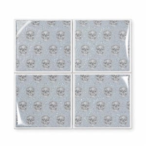 Peut inclure: Un ensemble de quatre dessous de verre carr&eacute;s pr&eacute;sentant un motif r&eacute;p&eacute;titif de cr&acirc;nes humains gris imprim&eacute;s sur un fond bleu clair avec une texture scintillante et paillet&eacute;e. Chaque dessous de verre a une bordure blanche nette.