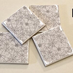 May include: A set of three square ceramic coasters featuring a glossy finish and a repeating black spider web pattern on a white background. The letter B is printed in black in the upper right corner of the image, indicating the product style.