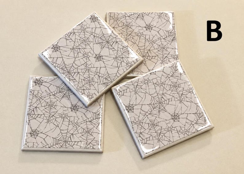 May include: A set of three square ceramic coasters featuring a glossy finish and a repeating black spider web pattern on a white background. The letter B is printed in black in the upper right corner of the image, indicating the product style.