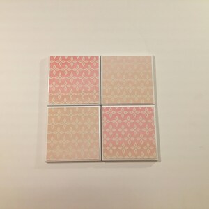 K&ouml;nnte beinhalten: Vier quadratische Keramikfliesen mit einem wei&szlig;en Blumenmuster auf rosa Hintergrund. Die Fliesen haben einen Verlaufseffekt, wobei die rosa Farbe zu einem helleren Rosaton verblasst.