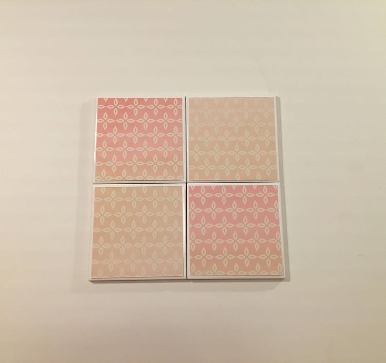 K&ouml;nnte beinhalten: Vier quadratische Keramikfliesen mit einem wei&szlig;en Blumenmuster auf rosa Hintergrund. Die Fliesen haben einen Verlaufseffekt, wobei die rosa Farbe zu einem helleren Rosaton verblasst.