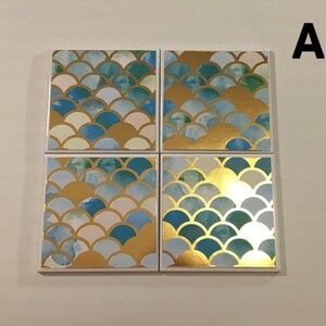 K&ouml;nnte beinhalten: Satz mit vier quadratischen Untersetzern mit einem blauen, wei&szlig;en und goldenen Schuppenmuster. Die Untersetzer sind aus Keramik und haben eine gl&auml;nzende Oberfl&auml;che.