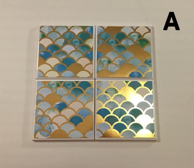 K&ouml;nnte beinhalten: Satz mit vier quadratischen Untersetzern mit einem blauen, wei&szlig;en und goldenen Schuppenmuster. Die Untersetzer sind aus Keramik und haben eine gl&auml;nzende Oberfl&auml;che.