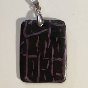 Puede incluir: Un colgante rectangular con una anilla de plata. El colgante est&aacute; hecho de arcilla polim&eacute;rica negra con un patr&oacute;n geom&eacute;trico morado.