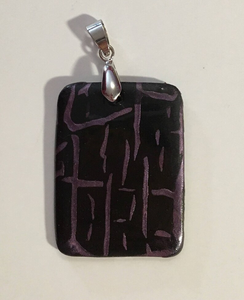 Puede incluir: Un colgante rectangular con una anilla de plata. El colgante est&aacute; hecho de arcilla polim&eacute;rica negra con un patr&oacute;n geom&eacute;trico morado.