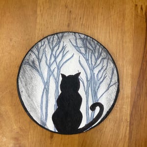 Pode incluir: Ilustra&ccedil;&atilde;o redonda a preto e branco com a silhueta de um gato preto em frente a uma cena de floresta. O gato est&aacute; de costas, com a cauda enrolada. O fundo mostra &aacute;rvores nuas numa paleta de cinza e branco nebuloso.