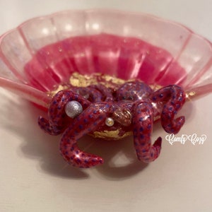 Puede incluir: Un plato de concha rosa con una figurita de pulpo p&uacute;rpura y rosa en su interior. El pulpo tiene detalles de purpurina dorada y cuentas de perlas.