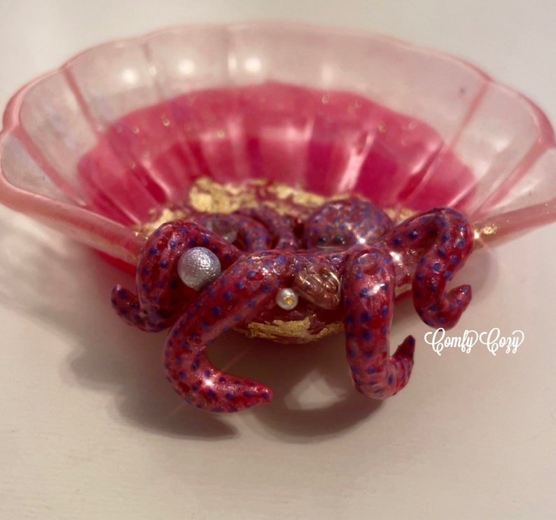 Puede incluir: Un plato de concha rosa con una figurita de pulpo p&uacute;rpura y rosa en su interior. El pulpo tiene detalles de purpurina dorada y cuentas de perlas.