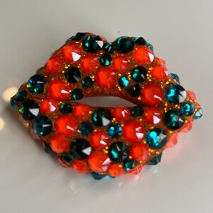 May include: A sparkling, jewel-encrusted lip-shaped brooch. The brooch features a brown base covered in vibrant orange and teal-colored rhinestones. The design is a bold and eye-catching accessory.