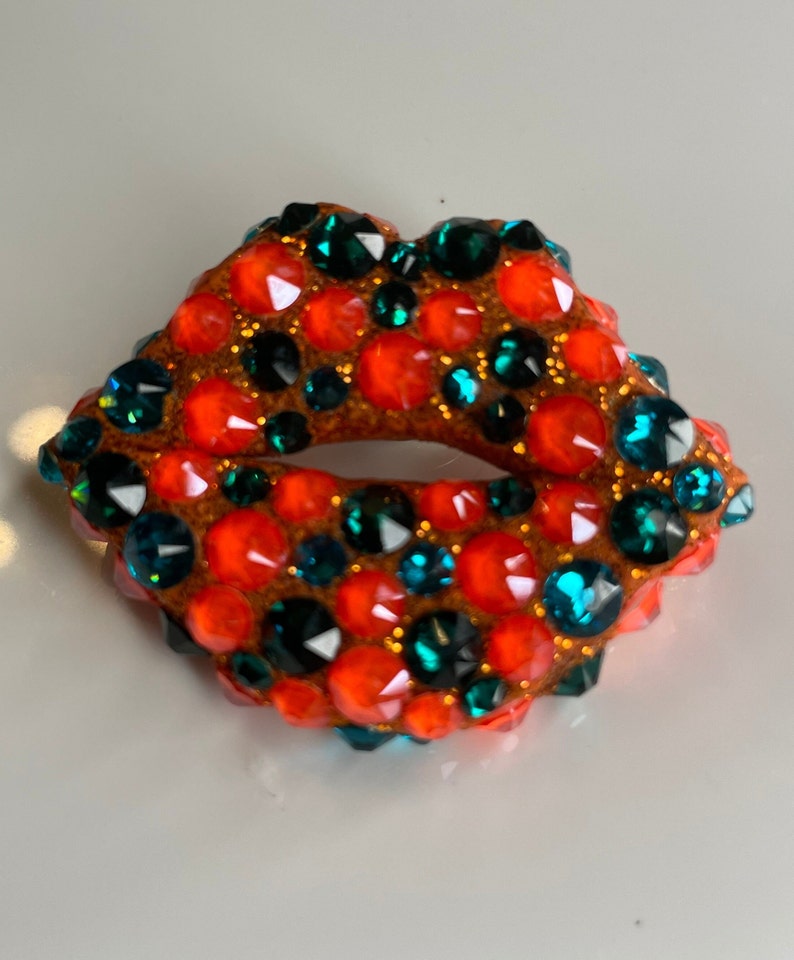 May include: A sparkling, jewel-encrusted lip-shaped brooch. The brooch features a brown base covered in vibrant orange and teal-colored rhinestones. The design is a bold and eye-catching accessory.