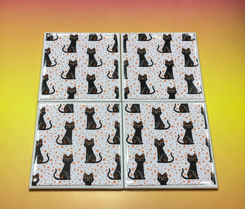 May include: A set of four square ceramic coasters featuring a repeating pattern of black cats with orange eyes and crescent moon markings on their foreheads, set against a white background filled with small orange stars and sparkles.